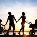 Family at sunset walking on the beach silhoutees are black with the sun setting behind them. Mum is pushing a push chair holding dads hands behind her and dads has his other hand reaching out to his som on his bike showing a united family