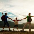 Mum, dad and child in the middle all with backpacks holding hands and looking out to the beautiful scenery lake and mountains