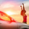 Woman throwing her hands up in the air by her car with sun setting behind her
