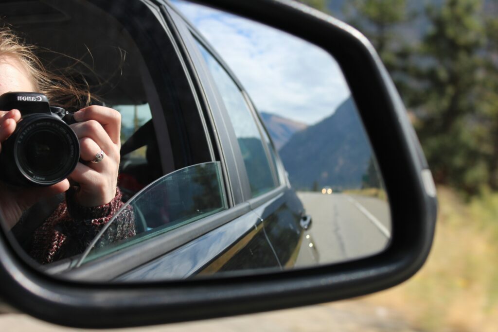 Reflection in a car's wing mirror of a girl taking a picture with her camera and the road and mountains