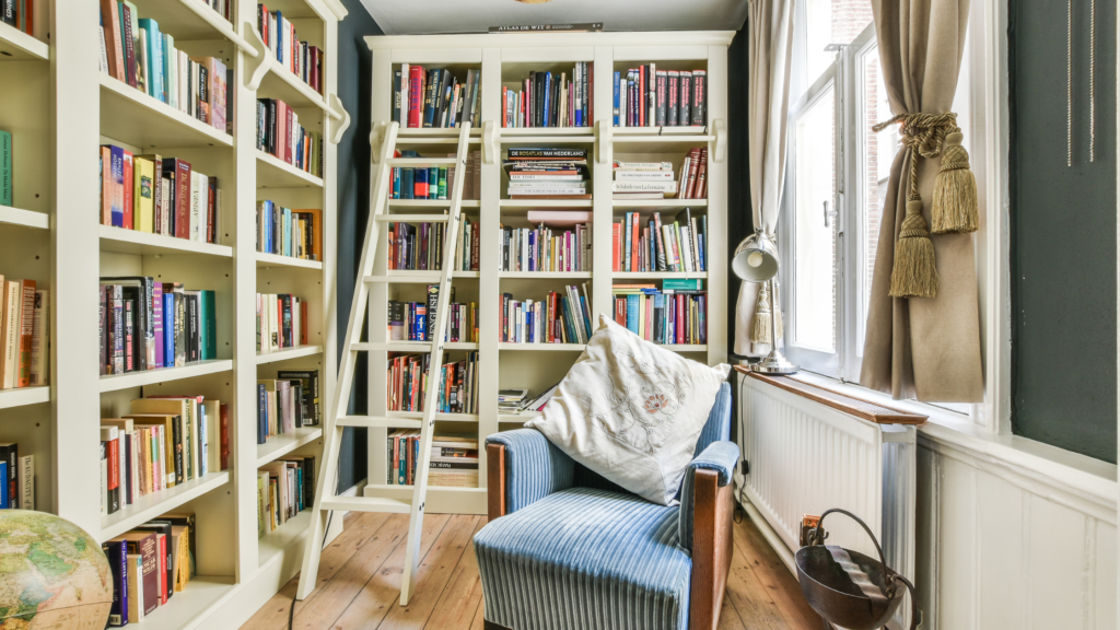Spare room set up as a library with floor to ceiling book shelves, a ladder to access high books and a blue comfy armchair with cushion