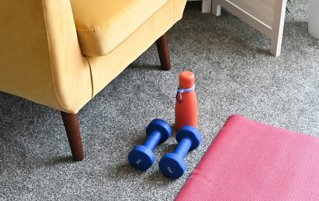 Some small blue weights, yoga mat and drink bottle on the living room floor with part of a yellow sofa