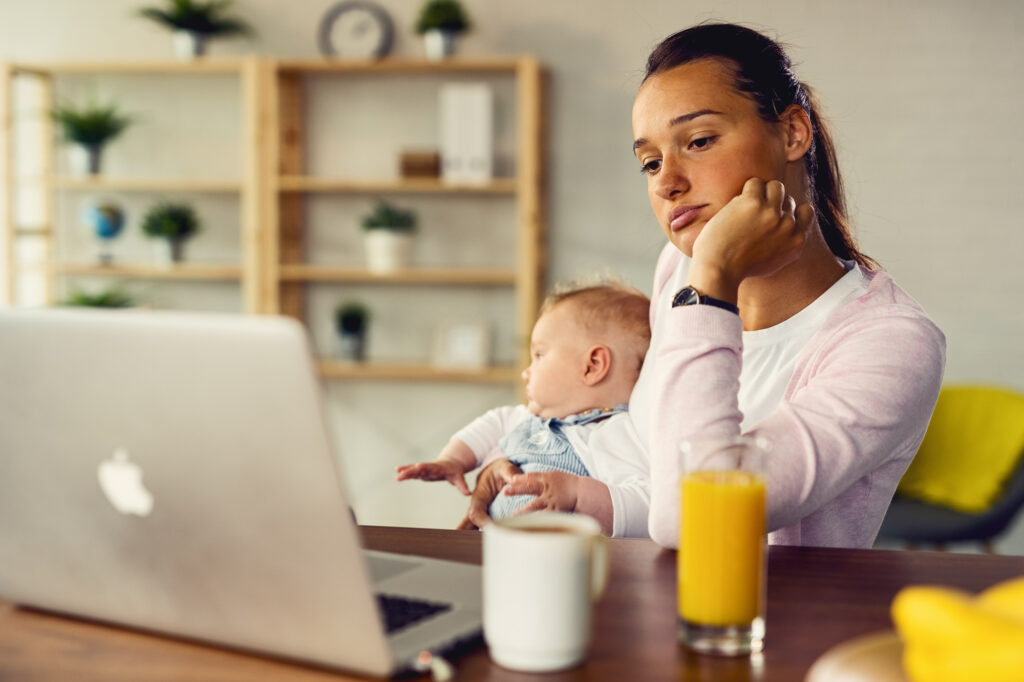 Tired mother working on her laptop while being with her baby at home.