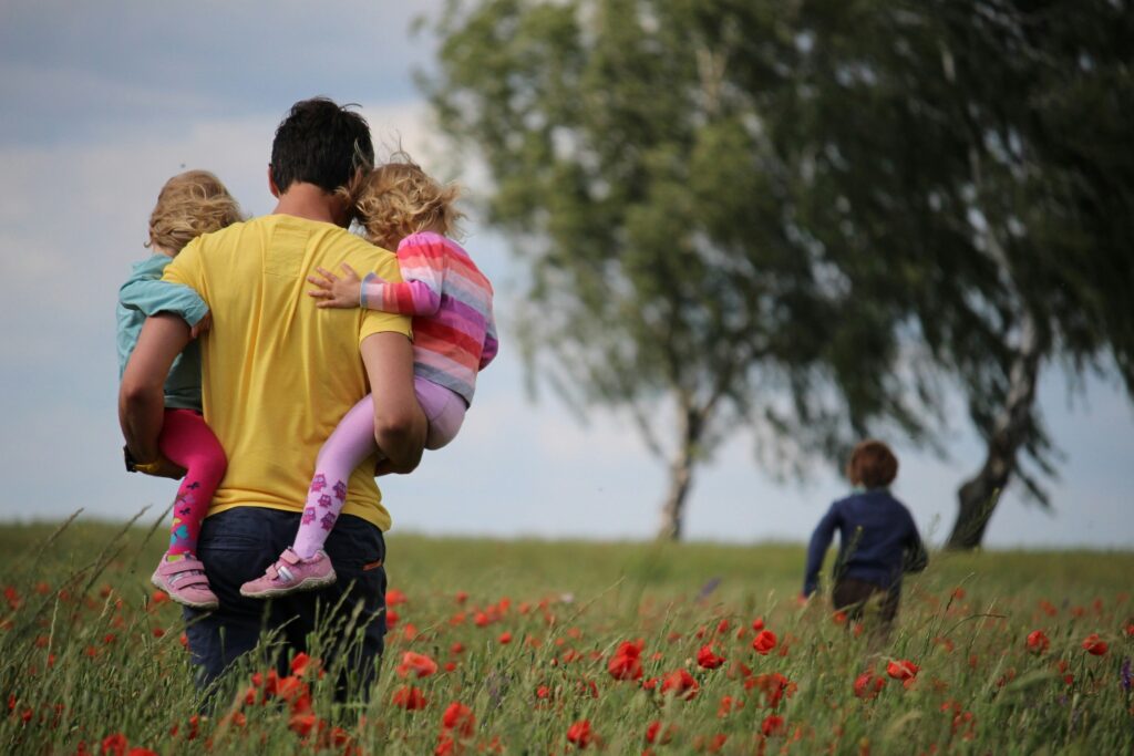 Dad carrying two children in his arms with one child running in front through a fielf of poppies. Picture taken from the back of the family.