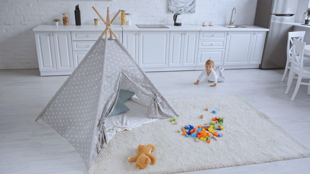 Toddler child in a kitchen living room with white sodr furnishings and cream berber rug with toys and a teepee set up on it.