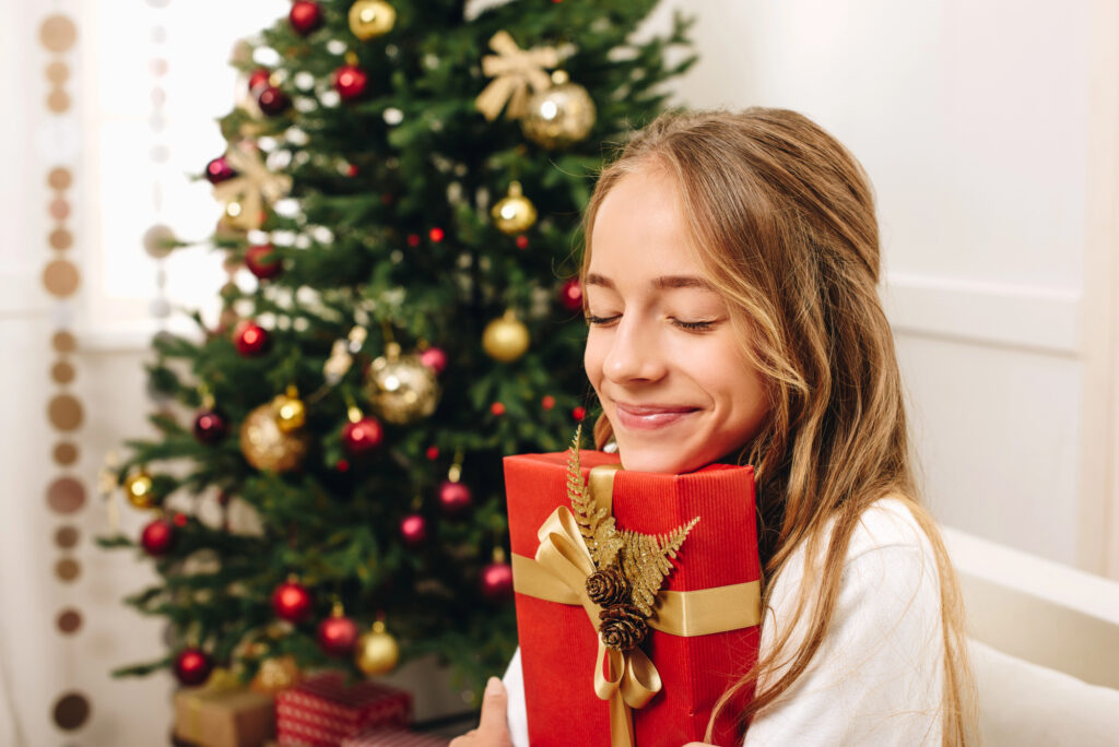 Teenage girl with wrapped Christmas present in red wrapping paper and gold bow in front of the Christmas tree looking happy.