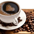 Coffee cup and saucer on a white background with coffee beans next to it on table. Coffee has heart shape.