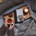 Cosy home with cup of herbal tea in female hands, plaid and candles surrounding her.