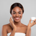 Skincare Routine. Beautiful Black Female Holding Jar With Moisturizing Cream And Applying On Face, Attractive African American Lady Nourishing Skin While Standing Wrapped In Towel On Grey Background