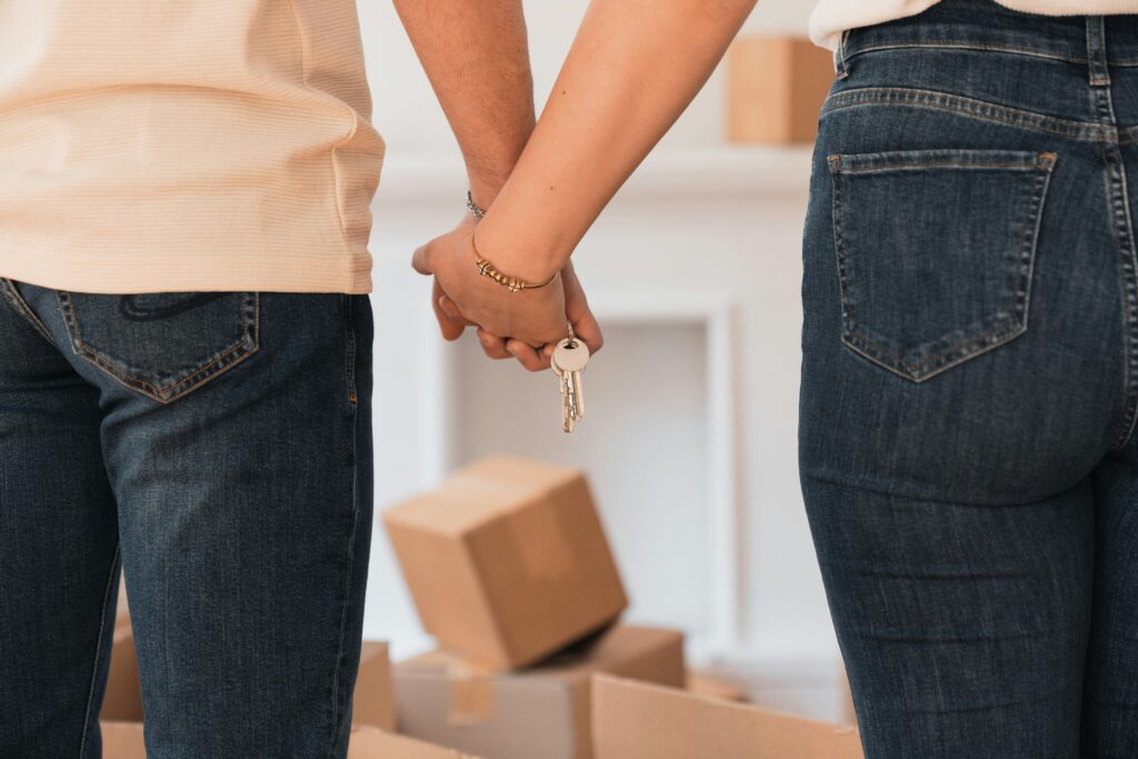 image from behind of the torsos of a couple holding hands with key between their hands and boxes in a room to represent moving to a new home.