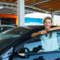 Happy looking woman smiling wityh keys in hand leaning against the door of a used car that she's just bought.