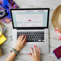 A desk laid out for travel planning with ladies hands on laptop with passport, summer hat and other travel essentials spread round.