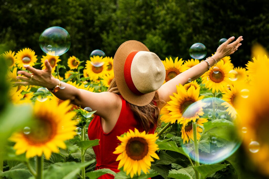 The back of a woman in a red dress and straw hat in a field full of sunflowers raising her arms in the air happily as bubbles float in the air.