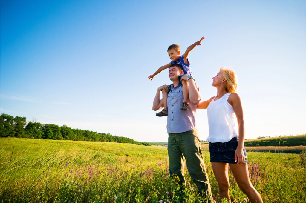 Mum and dad in a beautiful field together with their toddler on his father's shoulders pretending to be a plane
