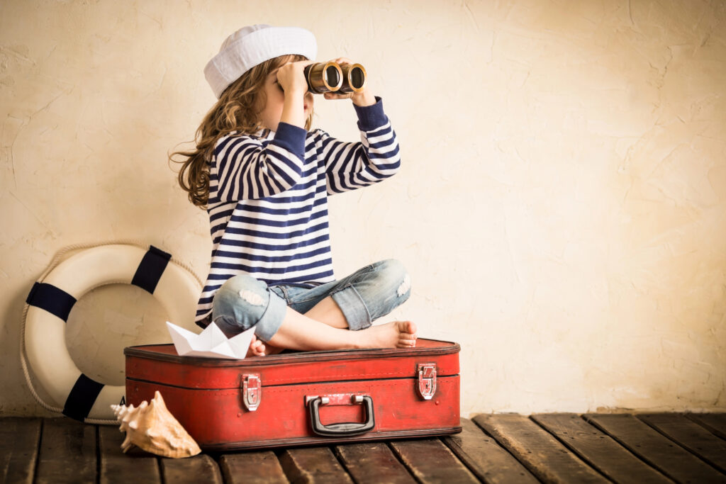 Young girl sitting on an old fashioned suitcase in stripe sailors top and captains hat, peering through binoculars