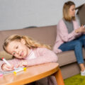 Young girl colouring in on her table with her mum in the background on the sofa.