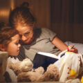 Mum putting child to be and reading a bedtime story snuggled up together with magical fairy lights