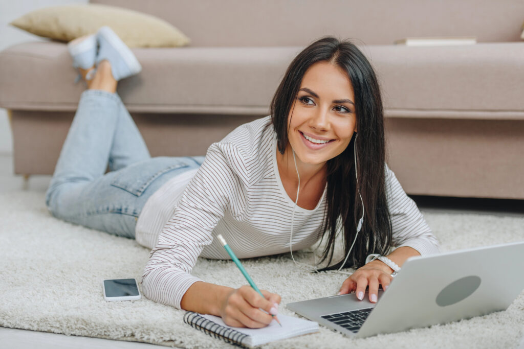 Young woman lying on the floor with her laptop and notepad and pen taking part in an online learning course