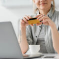 cropped view of young woman with tattoo on arm, holding a yellow credit card with coffee on her desk and facing her laptop