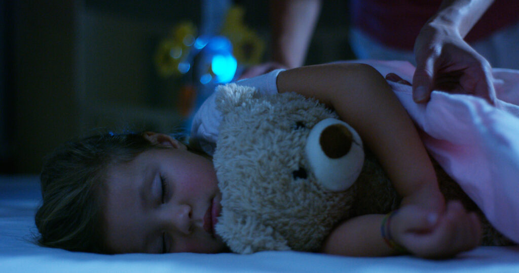 Young child being tucked in to sleep by her parent and cuddling her teddy bear in her bedroom with her eyes closed.