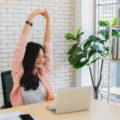 Female business owner stretching her arms up in the air at her desk facing her laptop in light and stylish office
