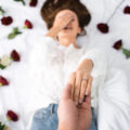 Smiling woman covering her face with one hand and holding her other hand up showing her engagement ring as she lies on a bed of roses.