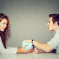 Couple sitting at the table opposite from each other tugging at a piggy banks looking angry and frustrated