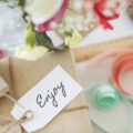 A wrapped parcel in brown packaging with gorgeous pastel ribbons on the table and a handwritten label saying 'enjoy'