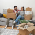 Couple sitting down on the sofa and smiling surrounded by boxes following their move to their new home