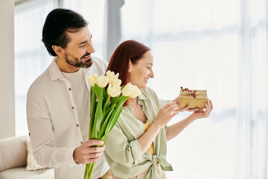 A couple with man holding flowers hugging his wife from behind whilst she looks excitedly at a wrapped gift